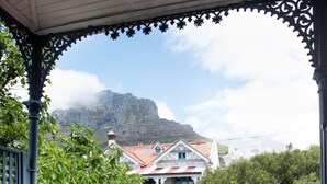 A balcony with two wooden chairs and a small table, overlooking a mountainous landscape and a building with a red roof.