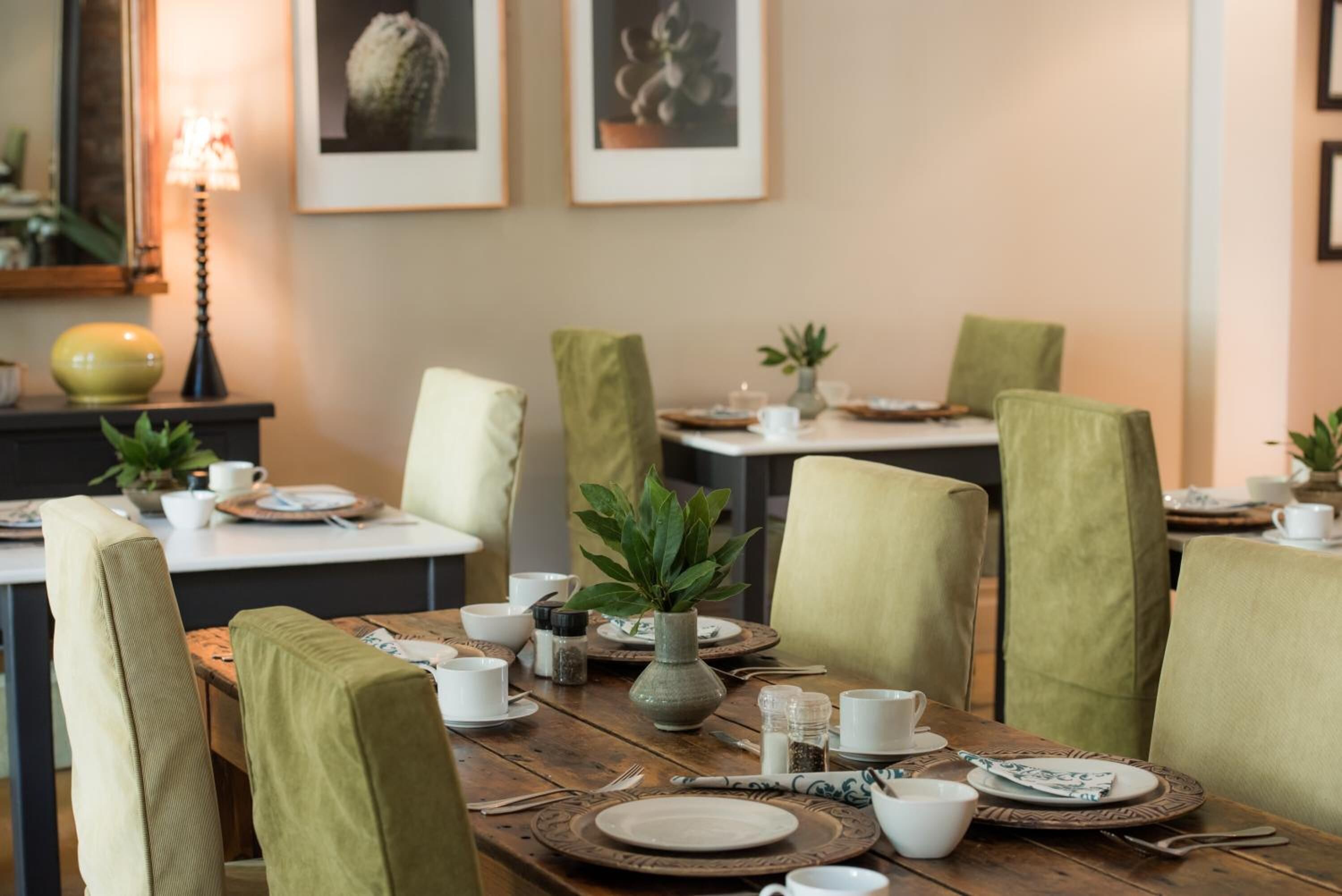 A dining area with wooden tables, green upholstered chairs, and a variety of table settings including plates, cups, and a vase with a plant.