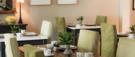 A dining area with wooden tables, green upholstered chairs, and a variety of table settings including plates, cups, and a vase with a plant.