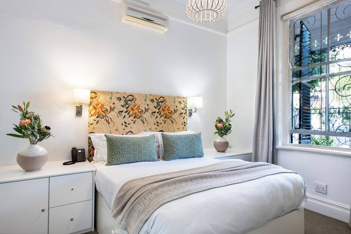 Un dormitorio ordenado con una cama, mesitas de noche, un teléfono y dos macetas.