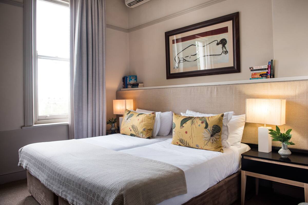 Una habitación de hotel con una cama grande, dos mesitas de noche, un cuadro enmarcado sobre la cama y una ventana con cortinas.