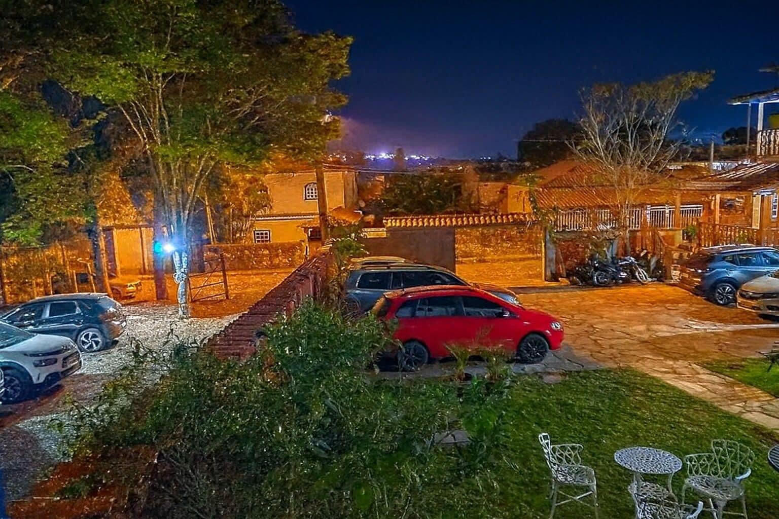 Un estacionamiento con varios coches, una pequeña zona de estar al aire libre con sillas y mesas de metal, y un edificio con tejado de baldosas al fondo.