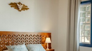 A bedroom with a wooden headboard, a bed with a light blue bedspread, two upholstered chairs, a nightstand with a lamp, and a window with curtains. - Pousada Ramalhete (Tiradentes)