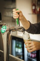 Una mano sostiene un vaso debajo de una llave de cerveza, con un refrigerador al fondo.