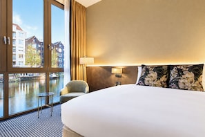 A hotel room with a large bed, a chair, a lamp, and a view of a canal with buildings. - Monet Garden Hotel Amsterdam (Amsterdam)