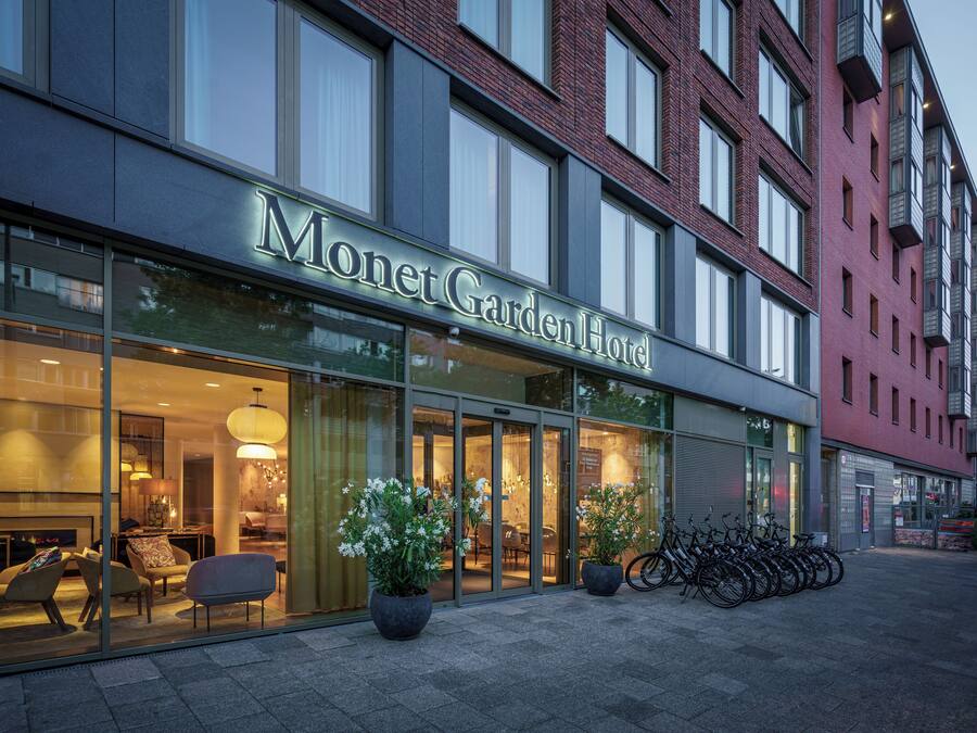 Exterior del Hotel Monet Garden con amplios ventanales, mobiliario exterior y plantas en macetas.