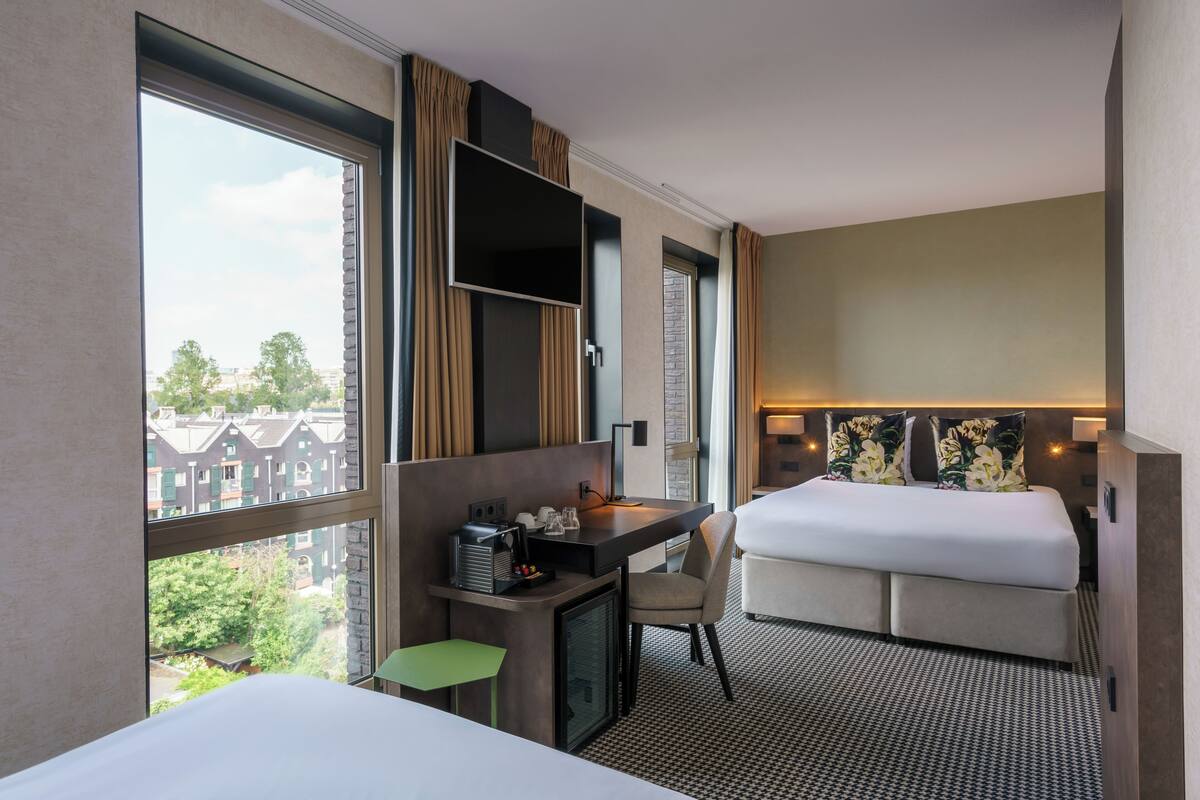 Ett hotellrum med en säng, ett skrivbord med en stol, ett litet kylskåp, en TV och ett stort fönster med gardiner.