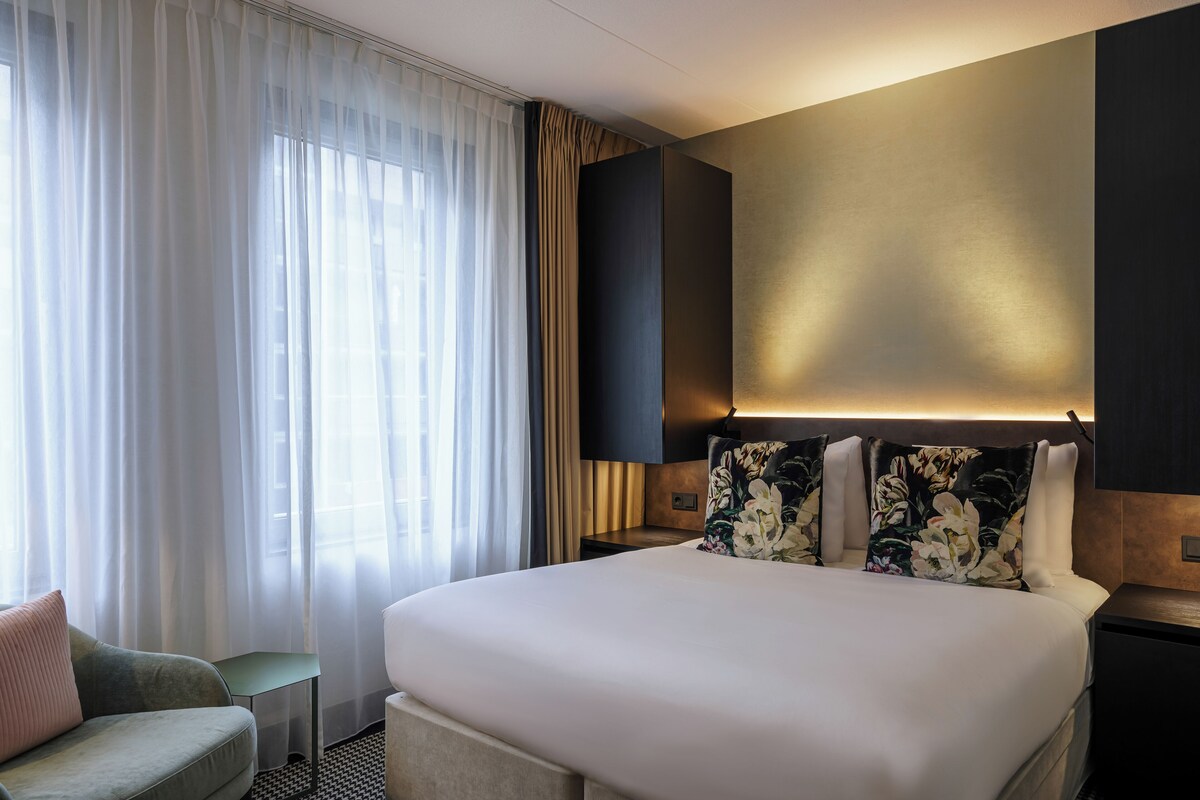 Ein Hotelzimmer mit einem großen Bett, einer kleinen Sitzecke und einem Fenster mit leichtem Vorhang.