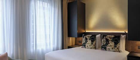 Quarto de hotel com uma cama grande, um espaço de estar pequeno e uma janela com cortinas translúcidas.