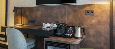 Una habitación de hotel moderna con una televisión de pantalla plana montada en la pared, un escritorio con lámpara, cafetera y un refrigerador pequeño.