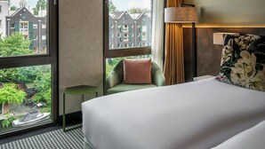Une chambre d’hôtel avec un grand lit, une vue sur les immeubles grâce à de grandes fenêtres, une petite table et une lampe.