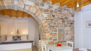A bedroom with a stone wall, a bed, a bedside table, and a small table with a plate of fruit. - Castle Mansions (Naxos)