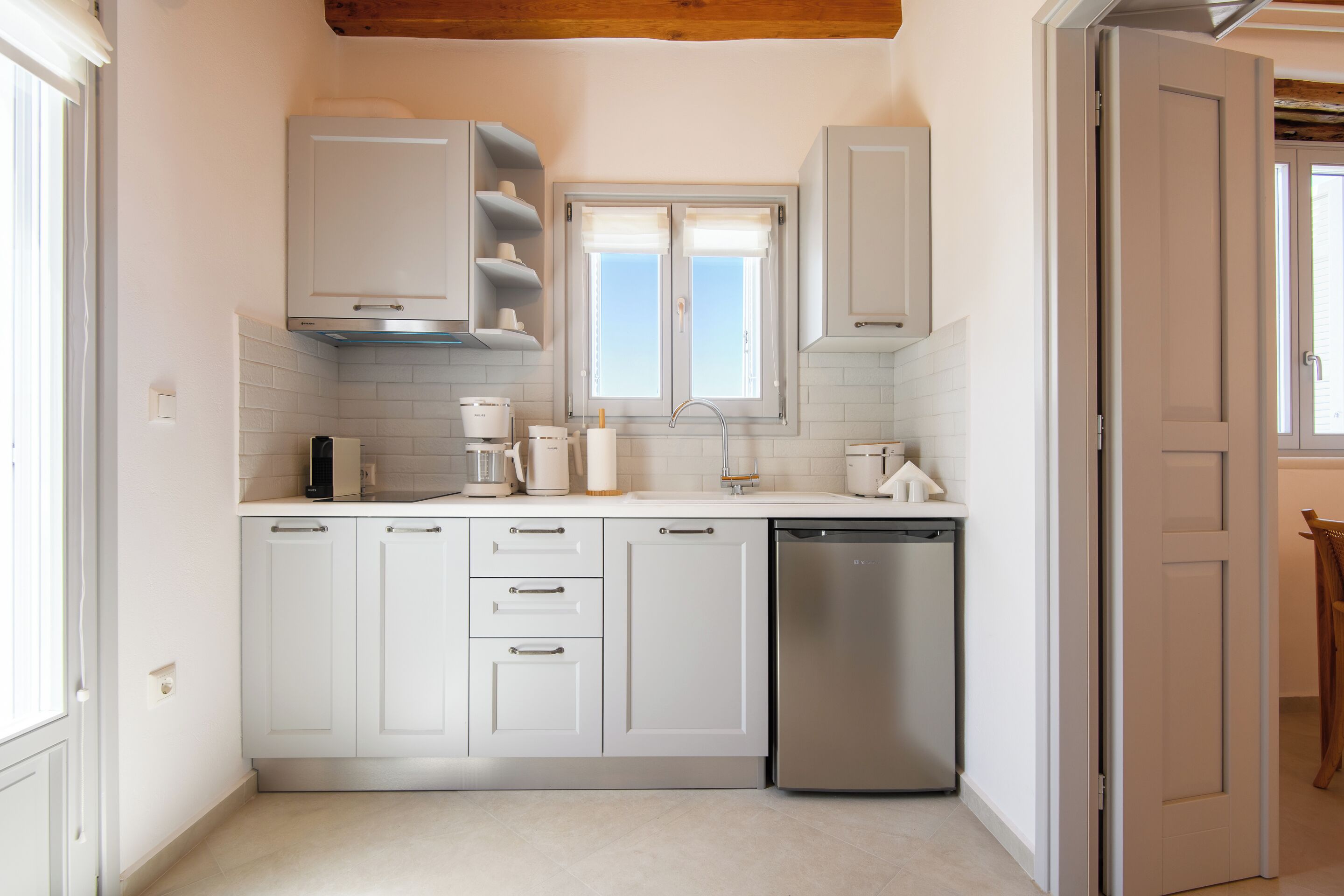 A compact kitchen with white cabinets, a stainless steel dishwasher, and a window with white curtains.