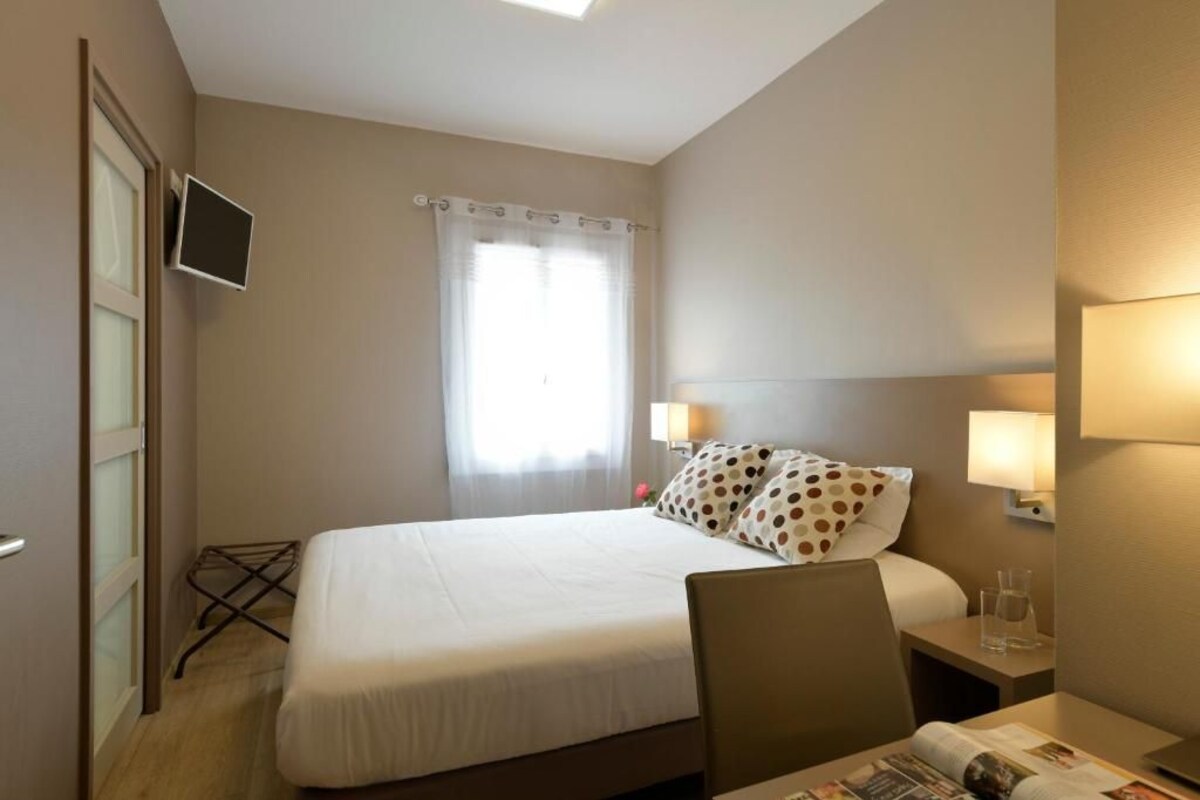 Habitación de hotel con cama, escritorio, silla, televisor montado en la pared y ventana con cortinas.