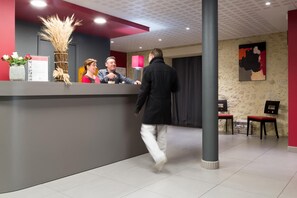 A reception area with a counter, a vase with dried plants, a lamp, and a painting on the wall. - Logis Hôtel Restaurant Le Pardaillan (Gondrin)