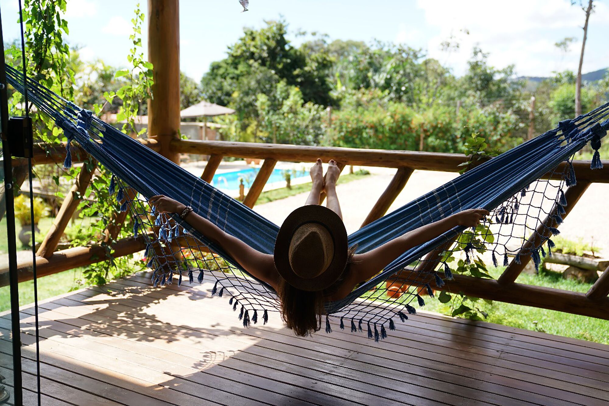 A hammock on a wooden deck with a view of a pool and greenery.
