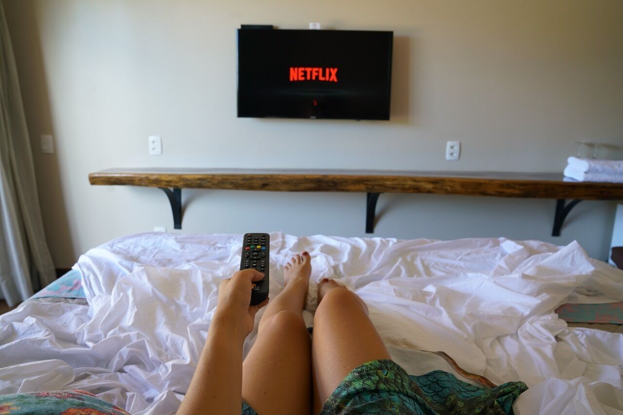 Uma cama com lençóis brancos, um banco de madeira e uma TV fixada na parede exibindo o logotipo da Netflix.