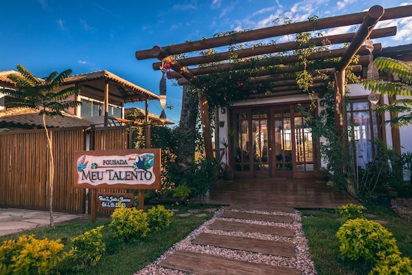 A wooden pergola entrance with a sign for 'Pousada Meu Talento' and a pathway leading to a building with glass doors.