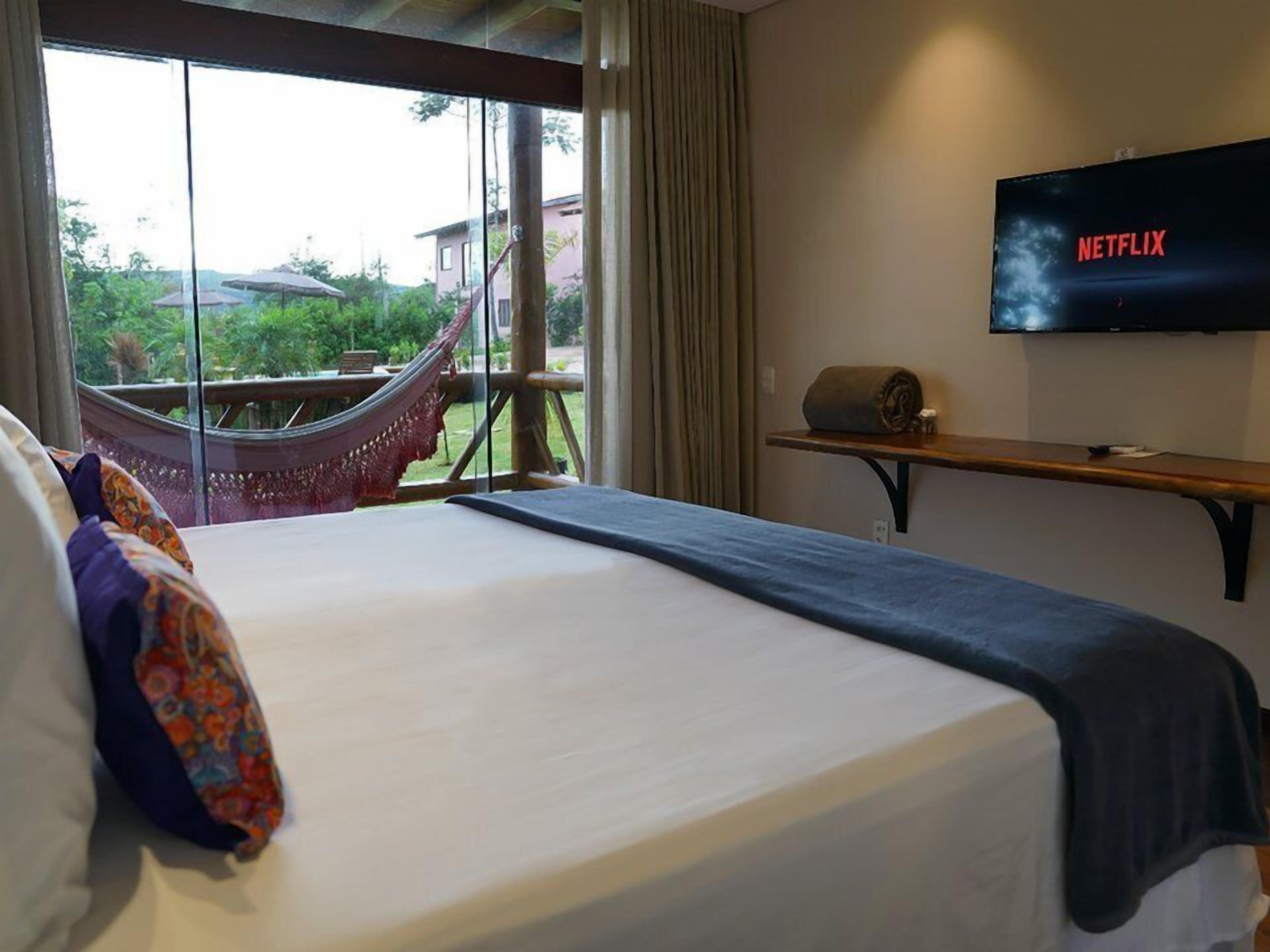 A hotel room with a large bed, a hammock, a TV on the wall, and a view of the outdoors.