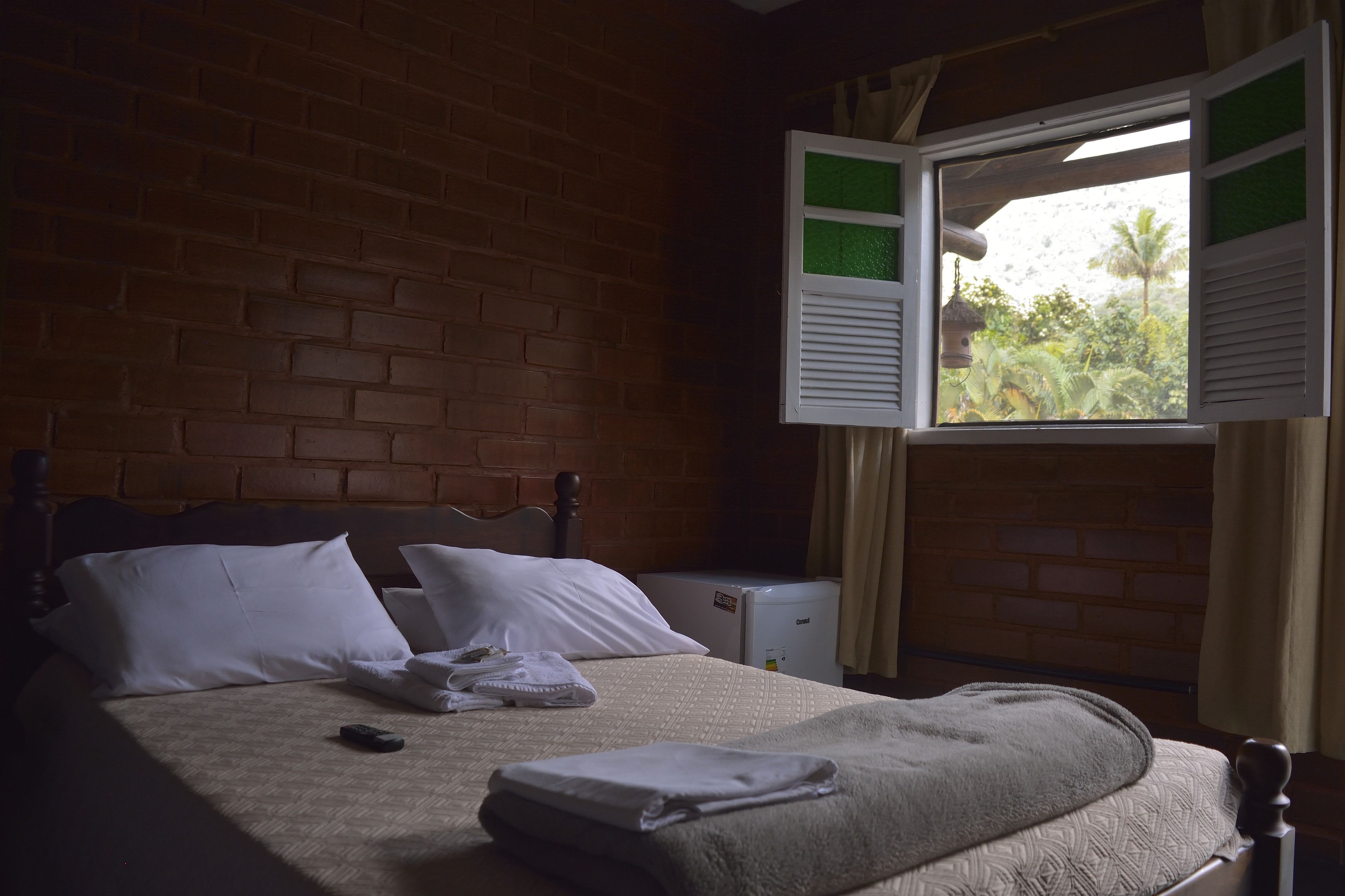 Een hotelkamer met een bakstenen muur, een bed met wit beddengoed, een klein nachtkastje en een raam met geopende luiken.