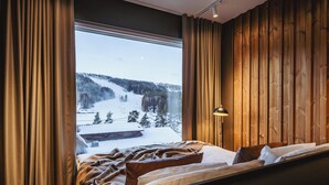 A bedroom with a large window, wooden panel walls, a bed with a blanket, and a bedside lamp.