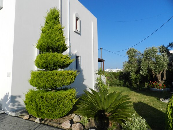 A white building with a series of green topiary bushes, a stone pathway, and a well-maintained lawn with various plants and a palm tree. - Triantafillas Appartments (Rhodes)
