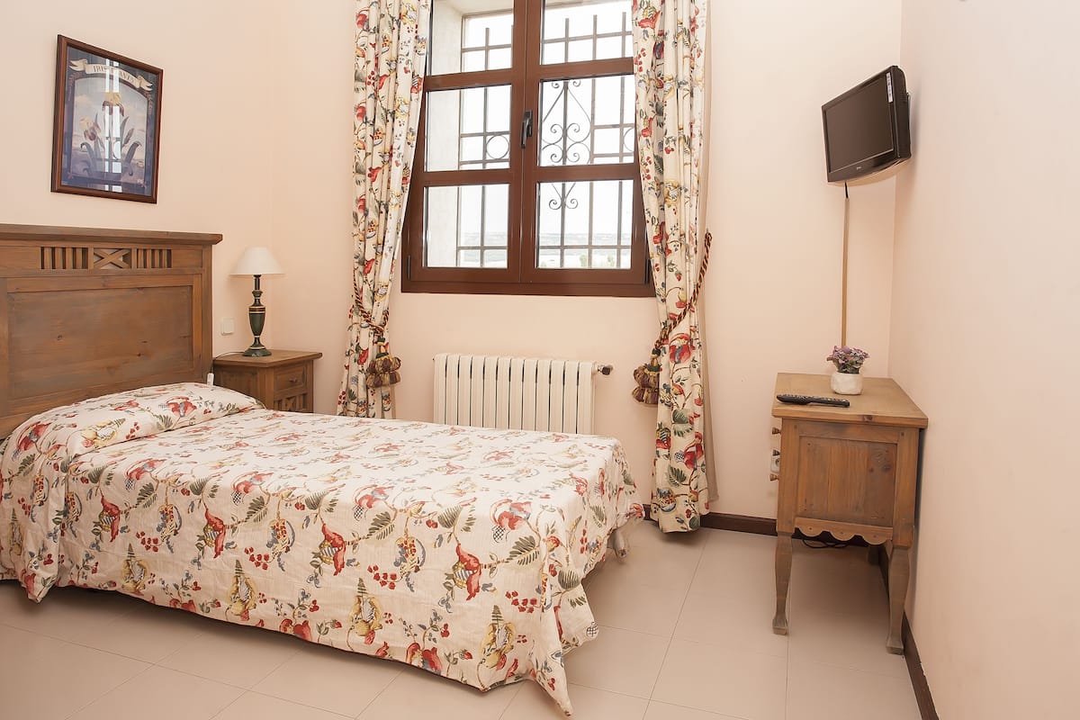 Un dormitorio con cabecera de madera, colcha con estampado floral, una mesita de noche con lámpara, un radiador, un televisor montado en la pared y una ventana con cortinas.