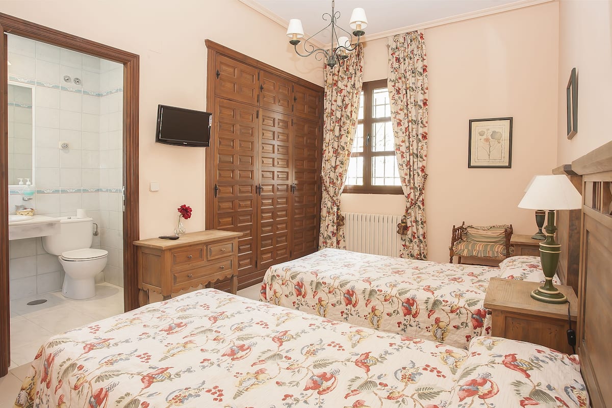 Un dormitorio con dos camas, un armario de madera, un televisor, una ventana con cortinas florales y una mesita de noche con lámpara.