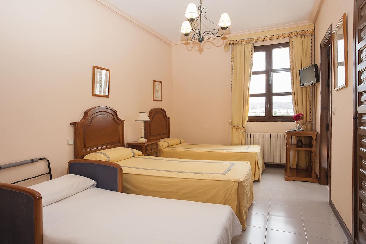 Habitación de hotel con dos camas individuales, una mesita de noche con lámpara, una pequeña mesa con un florero y una ventana con cortinas.