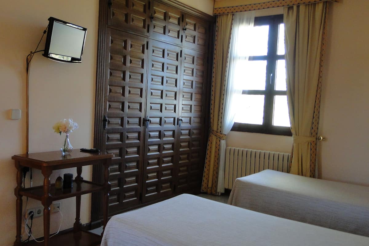 Habitación de hotel con un armario de madera, una mesita de noche con un jarrón de flores, una cama y una ventana con cortinas.