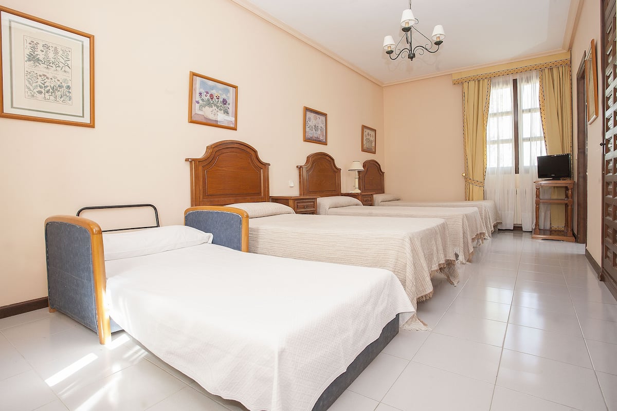 Una habitación de hotel con tres camas individuales, un escritorio con televisor y una estantería.