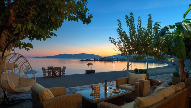 Eine Terrasse am Meer mit Korbmöbeln, einem Glastisch und Blick auf Wasser und Berge.