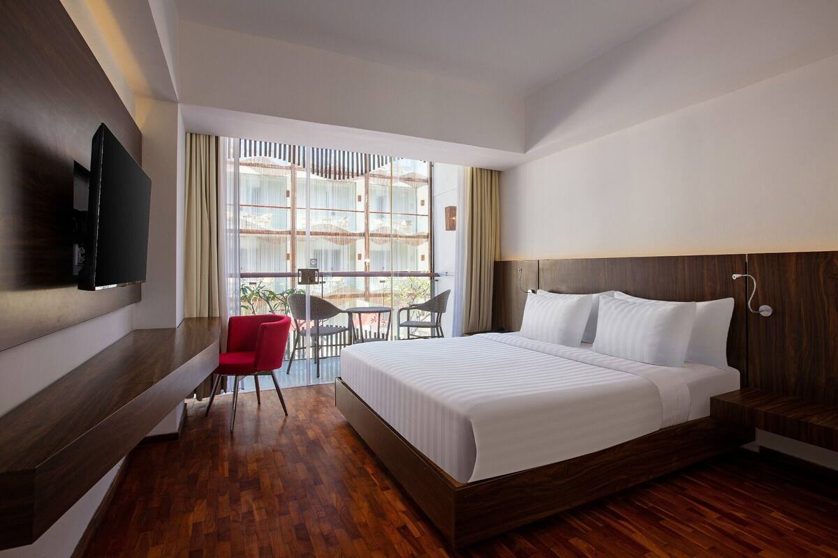 A modern hotel room with a large bed, a TV mounted on the wall, a small dining area with chairs, and a view through large windows.
