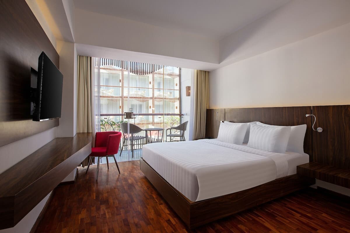 A modern hotel room with a large bed, a TV mounted on the wall, a small dining area with chairs, and a view through large windows.