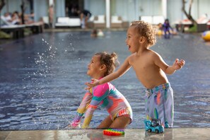 Deux enfants jouent dans une fontaine ou un bassin avec une petite voiture et un objet coloré qui se trouve par terre.