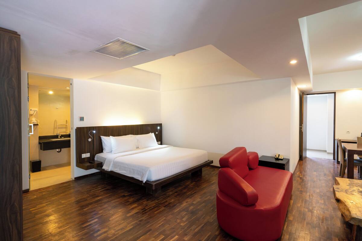 A modern hotel room with a wooden floor, a large bed with white linens, a red armchair, and a small wooden table.