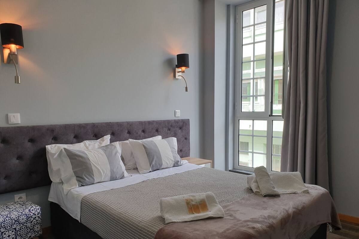 A neatly made bed with a grey headboard, two wall-mounted lamps, and a window with curtains.