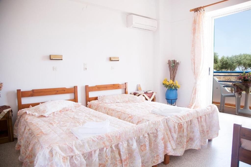 A bedroom with two beds, a window with a view, an air conditioning unit, and a small table with a vase.