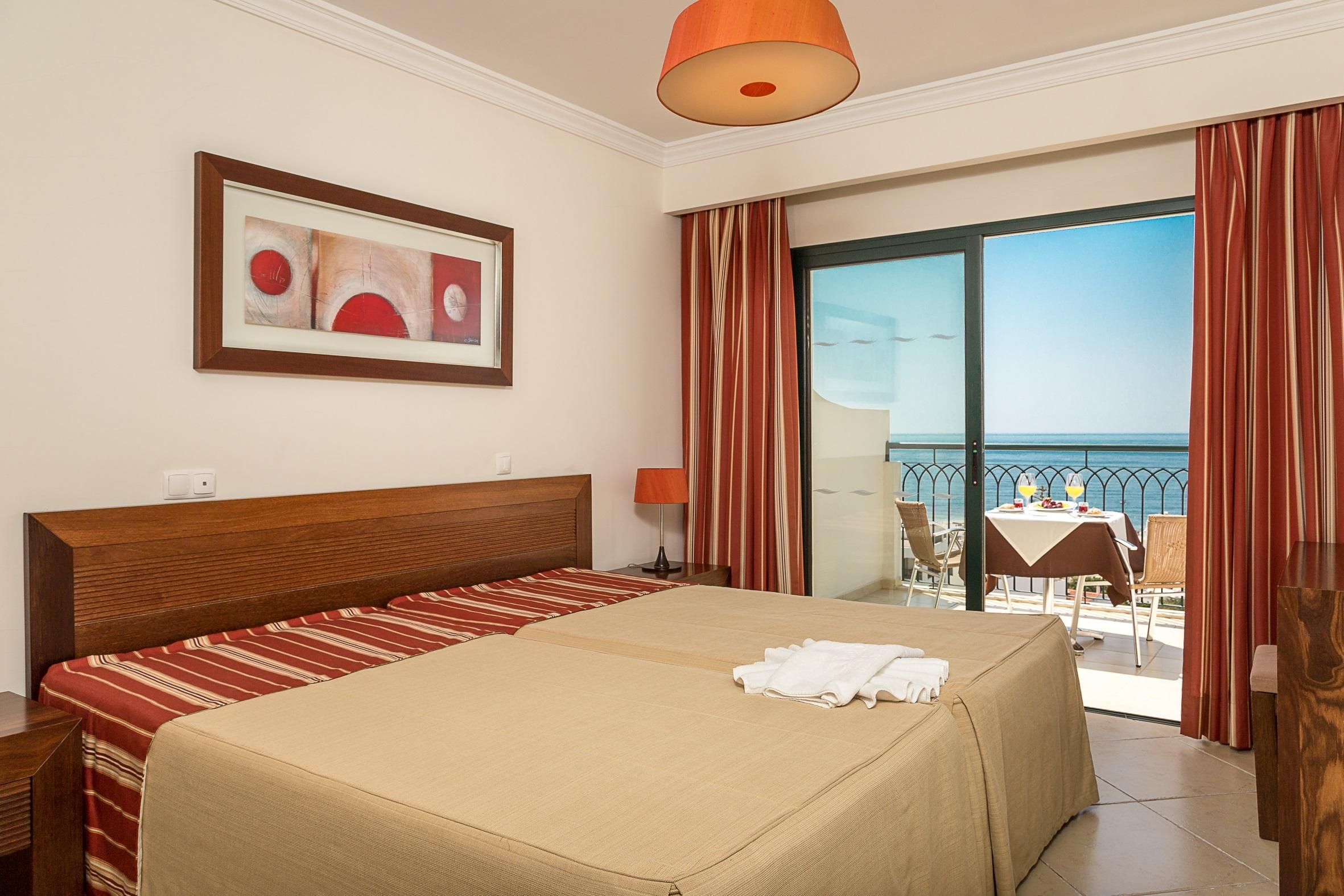 A hotel room with a bed, a painting, a table with chairs, and a view of the sea.