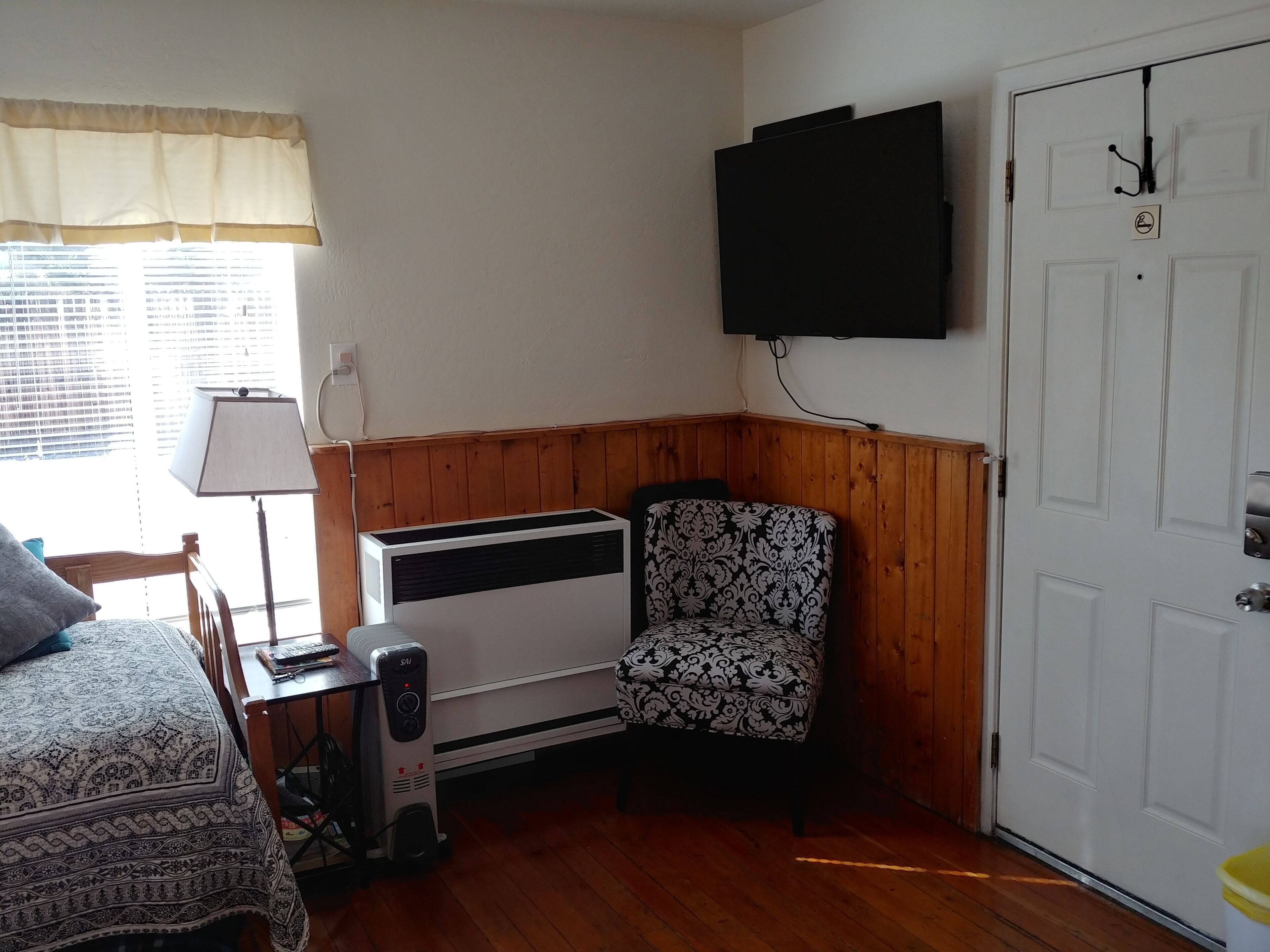 A room with a bed, a chair, a heater, a TV mounted on the wall, and a door.