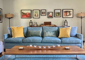 Una sala de estar con un sofá azul, cojines amarillos, una mesa de centro con vasos y cuadros en la pared.