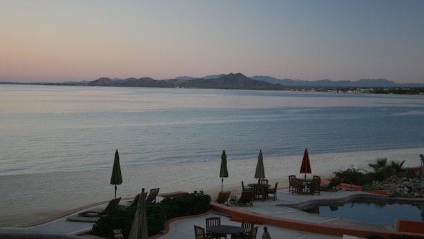 Zona de piscina en un resort con sillas de descanso y sombrillas, con vista al mar tranquilo y montañas a lo lejos.