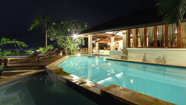 Ein Pool bei Nacht mit Liegestühlen, einem überdachten Bereich mit Vorhängen und tropischen Pflanzen.