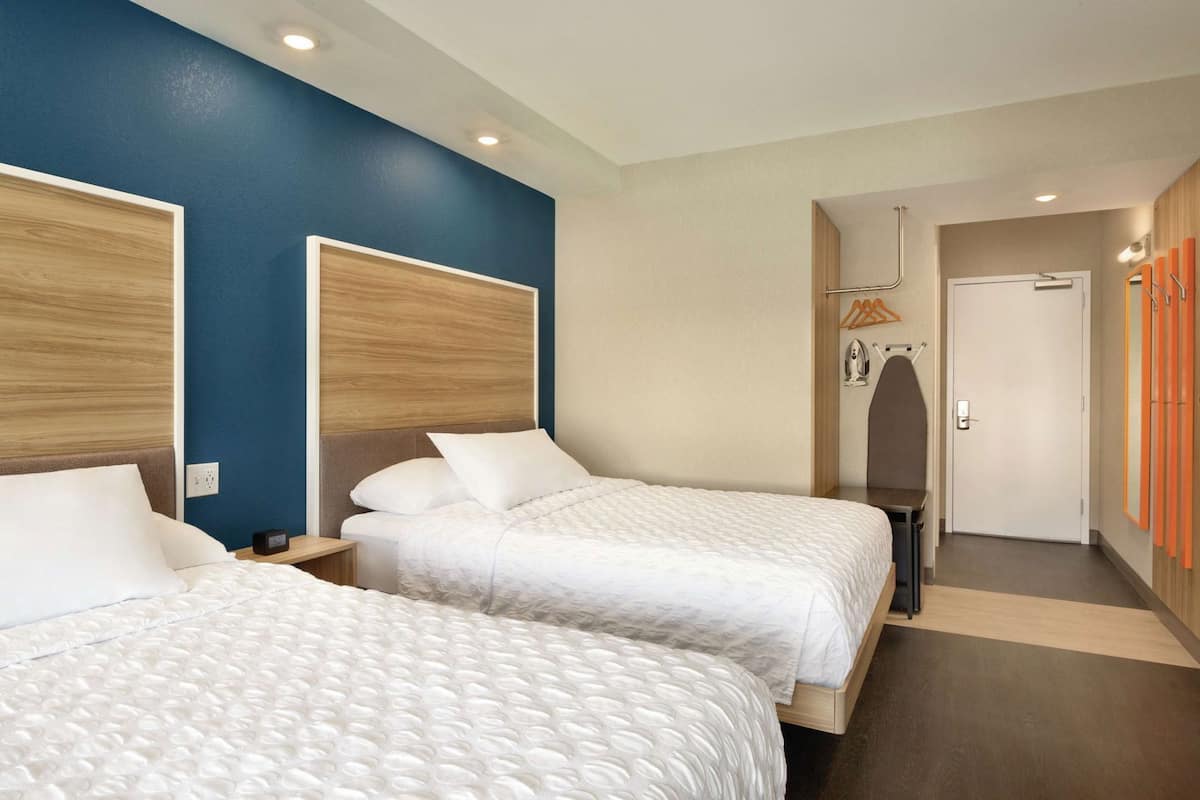 Ein Hotelzimmer mit zwei Betten, einem Holzkopfstück, einem Nachttisch, einem Kleiderschrank und einer Tür mit Kleiderhaken.