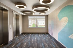 Un área de ascensor moderna con alfombra estampada, luces de techo circulares y un gran ventanal con vistas a edificios.