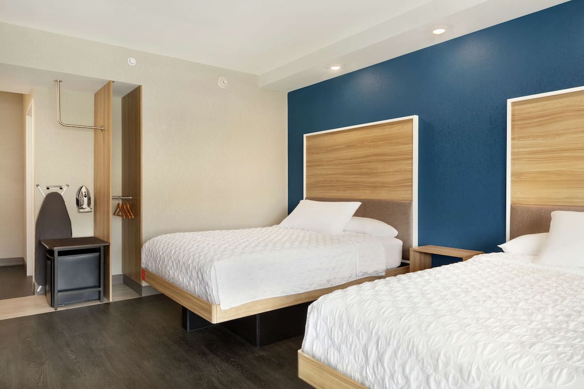 Ein Hotelzimmer mit zwei Betten, einem Holz-Kopfteil, einem kleinen Nachttisch, einem Spiegel und einem Kleiderschrank mit Kleiderstange.