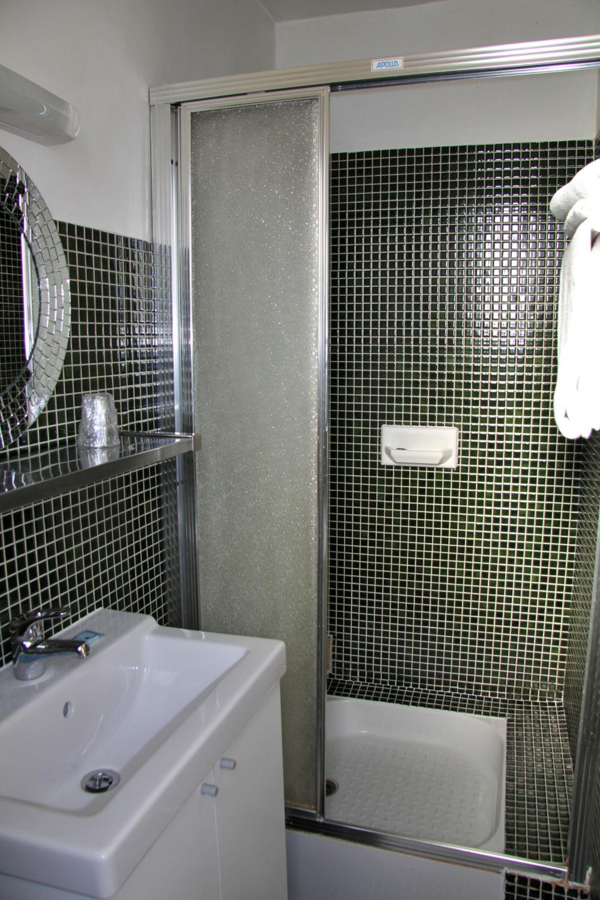 Un baño con lavamanos blanco, un espejo y una zona de ducha con pared de azulejos.