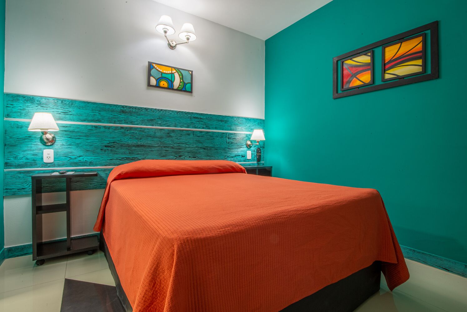 Una habitación con paredes color turquesa, una cama con colcha naranja, dos mesitas de noche con lámparas y dos lámparas fijadas a la pared.