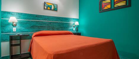 Una habitación con paredes color turquesa, una cama con colcha naranja, dos mesitas de noche con lámparas y dos lámparas fijadas a la pared.