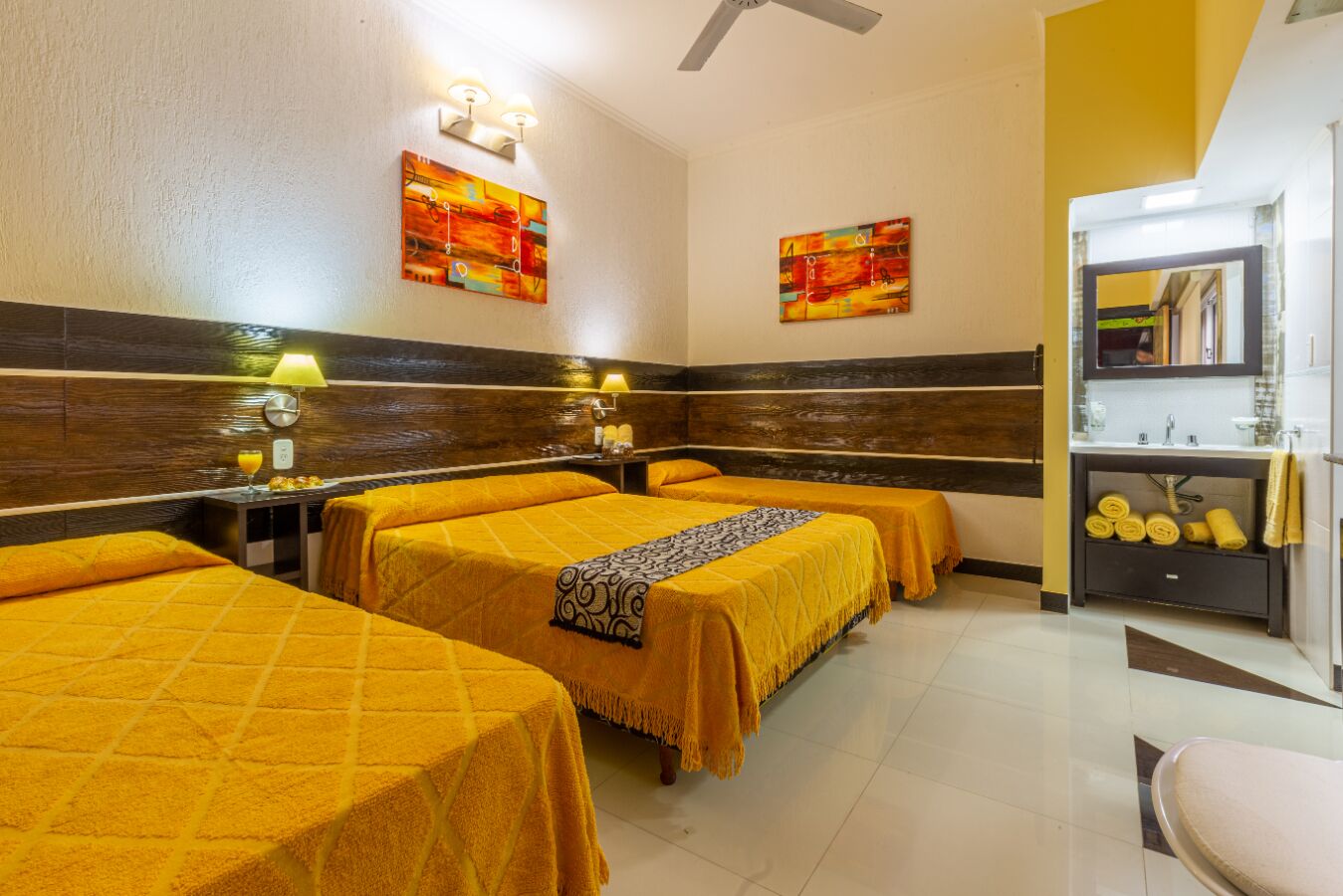 A hotel room with three beds, a bathroom with a mirror, and artwork on the walls.